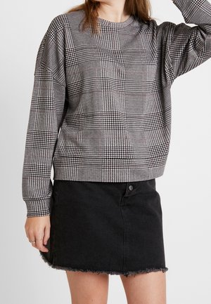 Femme portant un pull à manches longues pied-de-poule noir et blanc avec une mini-jupe en jean noir effilochée sur un fond uni.