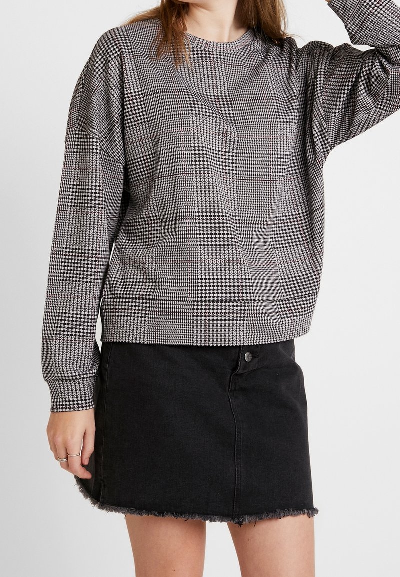 Femme portant un pull à manches longues pied-de-poule noir et blanc avec une mini-jupe en jean noir effilochée sur un fond uni.