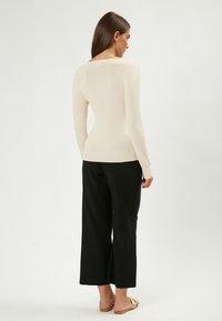 Top a manica lunga di un beige chiaro con design aderente, abbinato a pantaloni ampi neri e sandali flat, mostrato da dietro.