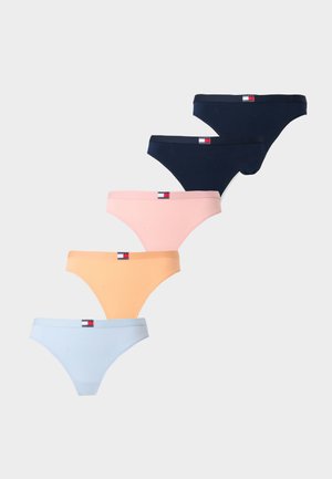 Ženska manekenka tamne puti s pletenicama stoji bosa, noseći bijeli Tommy Hilfiger bralette i tamnoplave gaćice, na jednobojnoj pozadini.