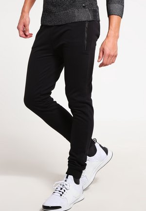 Mann trägt schwarze, schmal geschnittene Hose mit Reißverschlusstasche und weiße Turnschuhe, der auf einem einfarbigen Hintergrund einen Schritt nach vorne macht.