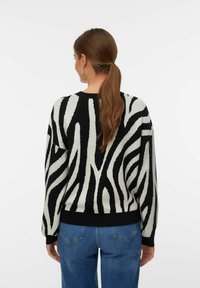Vrouw met bruin haar in een paardenstaart, gekleed in een zwart-wit gestreepte trui met zebramotief en blauwe spijkerbroek, staand tegen een effen witte achtergrond.