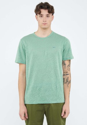 Giovane uomo con capelli ricci che indossa una maglietta verde chiaro a maniche corte e pantaloni color oliva, mostrando i tatuaggi sul braccio destro.