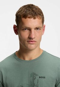 T-shirt di cotone verde con maniche corte, scollatura rotonda e stampa grafica con design geometrici; logo "BOSS" in nero.