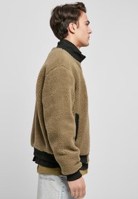 Rjava flisasta jakna z visokim ovratnikom, stranskimi žepi in črnimi poudarki. Mehka tekstura in prevelik kroj, nošena čez druge kose oblačil.