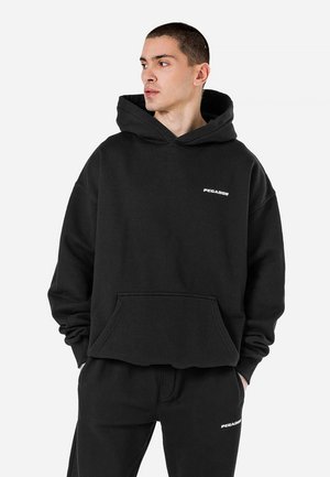 Hoodie noir en coton, avec une poche avant et le logo "PEGADOR". Coupe ample avec une grande capuche, accompagné de joggings assortis.
