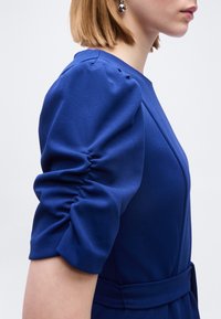 Γυναίκα με κοντά ξανθά μαλλιά που φοράει μπλε φόρεμα με ραβδωτά κοντά μανίκια και ασημένια σκουλαρίκια σφαίρα, εμφανιζόμενη από το πλάι σε επίπεδο κορμού.