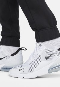 Vita Nike Air Max 270 sneakers med svarta swoosh-loggor, använda med vita strumpor och svarta uppvikta byxor på en vit bakgrund.