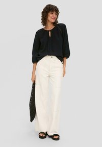 Czarna bluzka z długim rękawem z detalem w kształcie otworu, zestawiona z białymi spodniami o wysokim stanie i szerokimi nogawkami oraz czarnymi sandałami na platformie. Czarna torba typu tote.