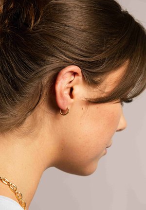 Goldene Creolenohrringe mit einer verdrehten Textur, die im Ohr einer Person getragen werden und das Profil ergänzen. Die Haare sind vom Gesicht wegfrisiert.