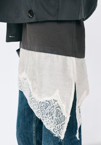 Dunkle Jacke und Hemd über einer mehrlagigen weißen Tunika mit Spitzenbesatz und seitlichem Schlitz, kombiniert mit blauen Denim-Jeans.