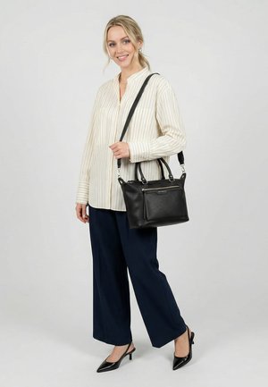 Femme portant un chemisier rayé et un pantalon bleu marine tient un sac à main en cuir noir à bandoulière, souriant devant un fond uni.