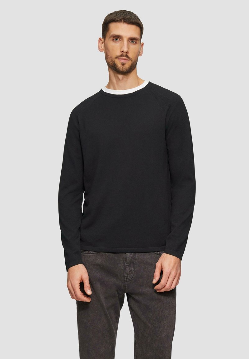 Zwarte trui met lange mouwen en ribbeltextuur, ronde hals en contrasterende witte kraag, gecombineerd met donkergrijze broek.