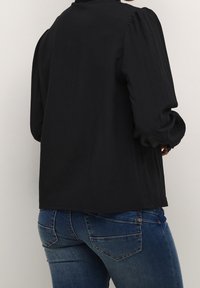Blouse noire à manches bouffantes, tissu texturé, coupe ample, dos légèrement plus long, portée avec un jean bleu foncé doté de poches arrière.