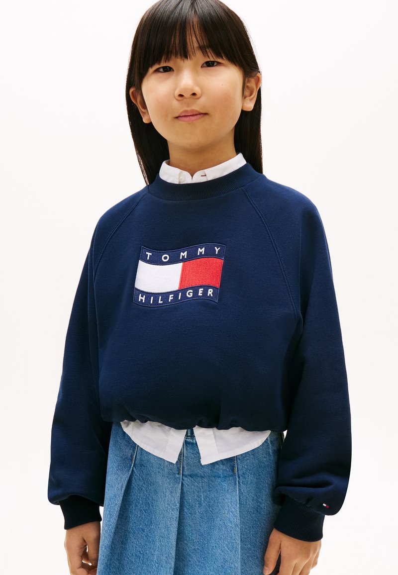 Νεαρή κοπέλα που φοράει μπλε μπλούζα Tommy Hilfiger πάνω από λευκό πουκάμισο και μπλε τζιν φούστα, στέκεται μπροστά σε λευκό φόντο.