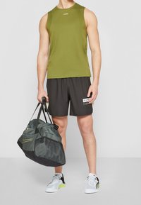 Haut sans manches vert olive, short noir avec des touches blanches et un logo, tenant un sac de sport vert foncé avec des détails en mesh, baskets blanches et noires.