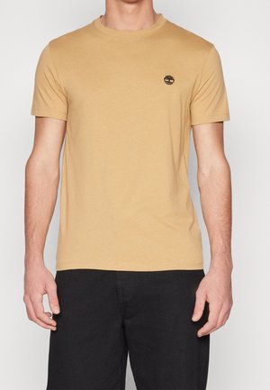 Mann trägt ein schlichtes beiges T-Shirt mit kurzen Ärmeln und einem kleinen schwarzen Baum-Logo auf der linken Brust, kombiniert mit schwarzen Hosen.