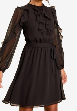 Femme portant une robe noire longueur genou avec manches longues transparentes, détails à volants sur le devant et une ceinture nouée à la taille.