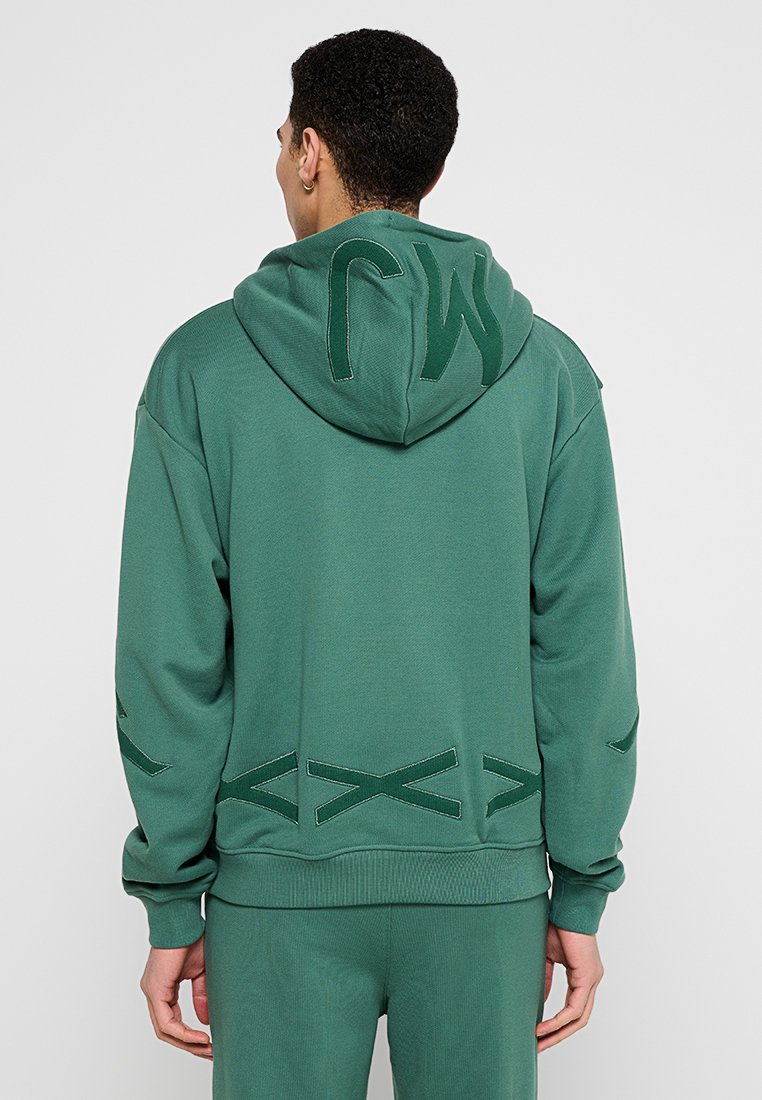 Homme portant un sweat à capuche vert et un pantalon assorti, de dos visible, avec des lettres brodées sur la capuche et des motifs géométriques sur les manches et la taille.