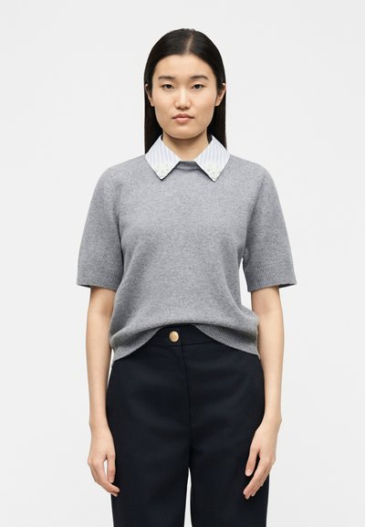 Femme aux longs cheveux foncés portant un pull gris à manches courtes sur une chemise rayée à col, et un pantalon noir, debout devant un fond blanc.