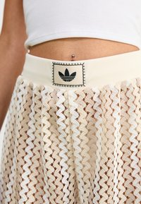 Kermakoshoit, joissa on sahalaitakuosi, paksu joustava vyötärö, johon on kiinnitetty musta Adidas-logo neliömäisellä tarralla.