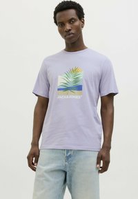 Camiseta de algodón lila de manga corta, con un gráfico de hoja tropical y logo en blanco. Combinada con unos jeans de mezclilla azul claro.