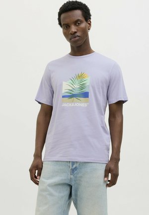 Camiseta de algodón lila de manga corta, con un gráfico de hoja tropical y logo en blanco. Combinada con unos jeans de mezclilla azul claro.