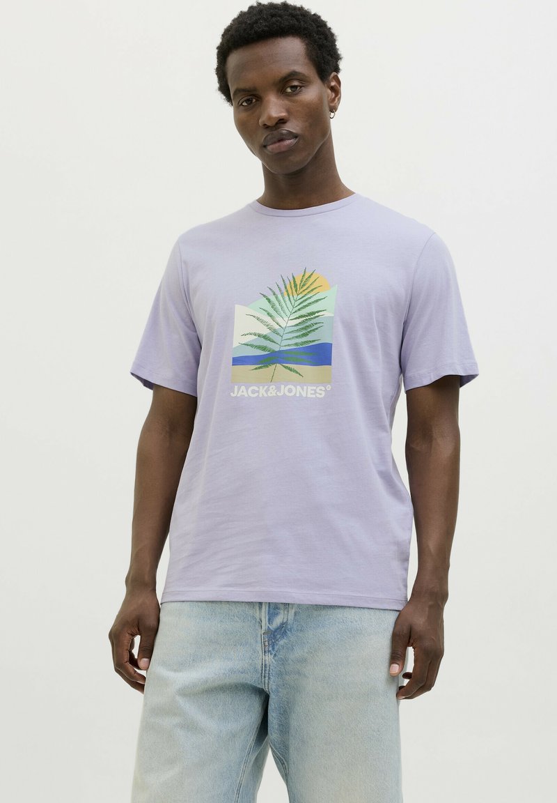 Camiseta de algodón lila de manga corta, con un gráfico de hoja tropical y logo en blanco. Combinada con unos jeans de mezclilla azul claro.