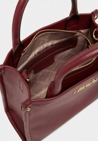 Sac à main en cuir bordeaux avec deux poignées, fermeture éclair et poches intérieures. Équipé de quincaillerie dorée et d'une doublure imprimée du logo.