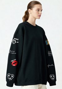 Mladá žena v černé oversized mikině se barevnými nášivkami a textem na rukávech, v kombinaci s kalhotami světlé barvy, dívající se doprava.