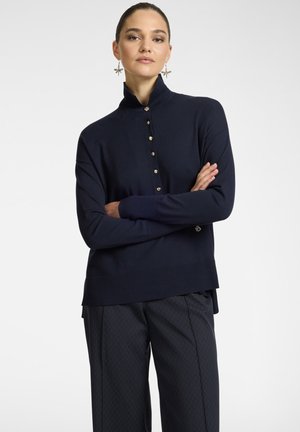 Maglione a maglia di colore navy con colletto alto e una chiusura a sei bottoni. Presenta polsini e orlo a coste, abbinato a pantaloni scuri con motivo.