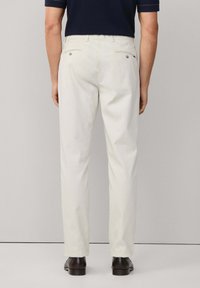 Muškarac u bež hlačama, tamnosmeškim poslovnim cipelama i mornarskoplavoj majici kratkih rukava, stoji uz jednostavnu svetlosivu pozadinu.