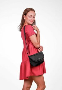 Černá křížová taška zaobleného tvaru, se zipovým uzávěrem a detailem loga. Modelka má na sobě červené šaty s rozšířenou sukní. Jednoduchý design.