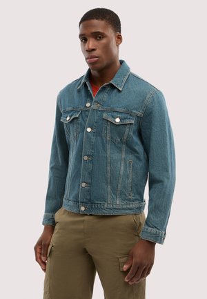 Giovane uomo che indossa una giacca di jeans blu con bottoni anteriori e pantaloni cargo kaki, in piedi davanti a uno sfondo chiaro e uniforme.