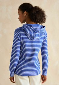 Žena s kučeravými vlasmi, oblečená v modro-bielej pruhovanej mikine, zobrazená zozadu, stojaca proti neutrálnemu pozadiu.
