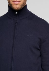 Marinblå zip-up tröja med hög krage, ribbad textur. Har en diskret logotyp på bröstet och en slät dragkedja.