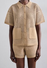 Strikket todelt sæt i beige med åbent diamantmønster, der består af en kortærmet top med krave, knapper og to forlommer foran.