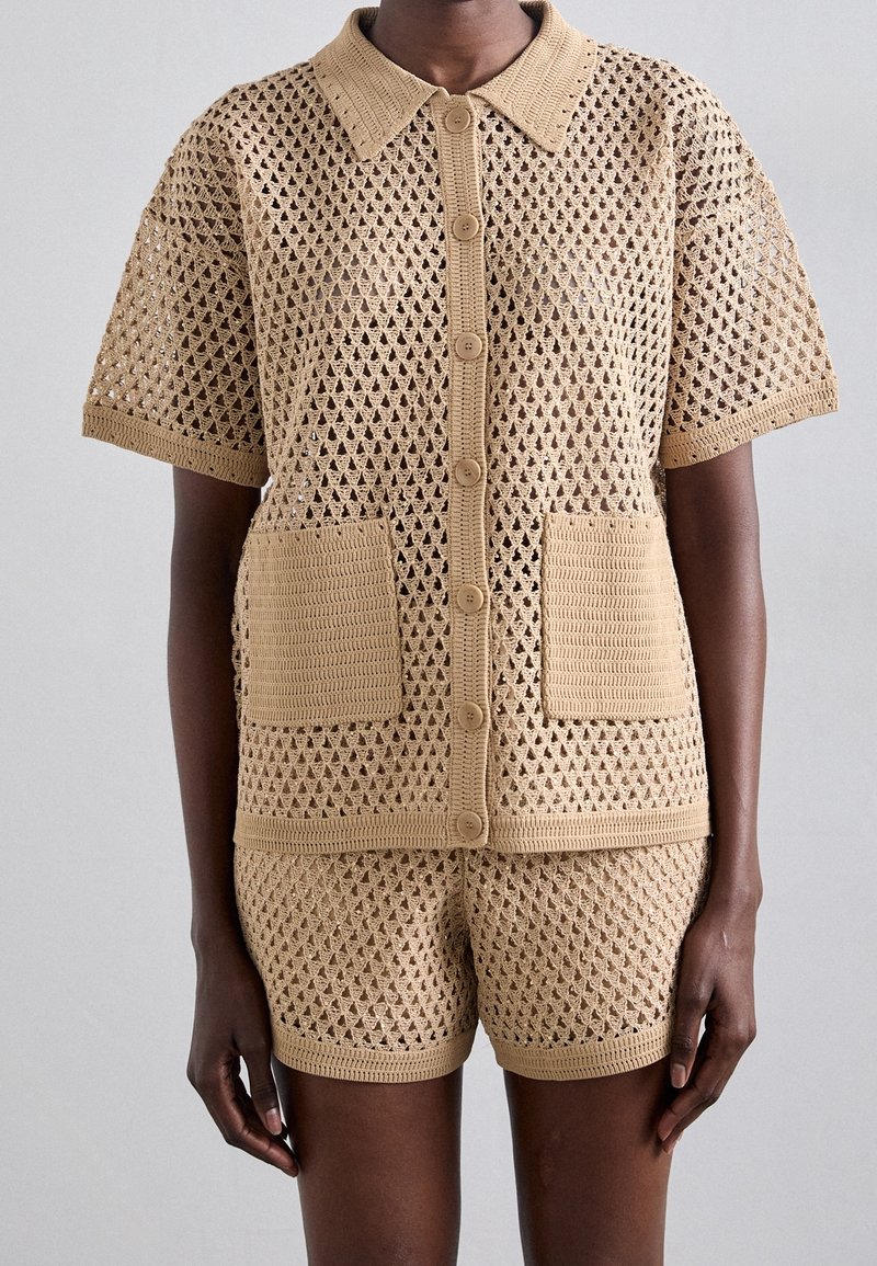 Strikket todelt sæt i beige med åbent diamantmønster, der består af en kortærmet top med krave, knapper og to forlommer foran.