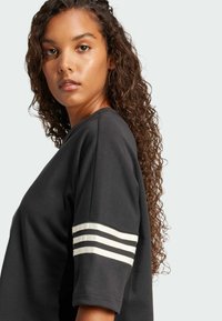 Čierne oversize tričko s okrúhlym výstrihom a tromi bielymi pruhmi na manžetách rukávov. Hladká bavlnená tkanina a voľný strih.