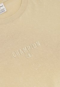 Beige katoen T-shirt met een geribde ronde hals. Heeft een subtiel, geborduurd "CHAMPION" logo op de borst en een lichte textuur in de stof.