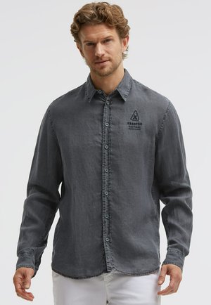 Mann trägt ein verblasstes dunkelgraues Hemd mit Knöpfen und Logo auf der Brust sowie weiße Hosen, steht vor einem schlichten hellen Hintergrund.