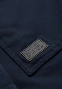 Tissu bleu marine avec une étiquette en cuir noir texturé, affichant un texte en relief : « OMBRE WEAR DEPUIS 1985 NOUVEAU DESIGN ». Bords cousus visibles.