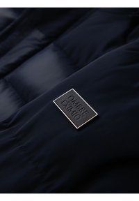 Acercamiento a una chaqueta acolchada de color azul marino con una etiqueta rectangular de la marca "OMBRE EXIMIO" en negro y plata.