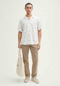 Korte mouwen, wit bloemenhemde met lichtblauwe en groene patronen, gecombineerd met beige broeken en crèmekleurige sneakers. Houdt een beige tote bag vast.