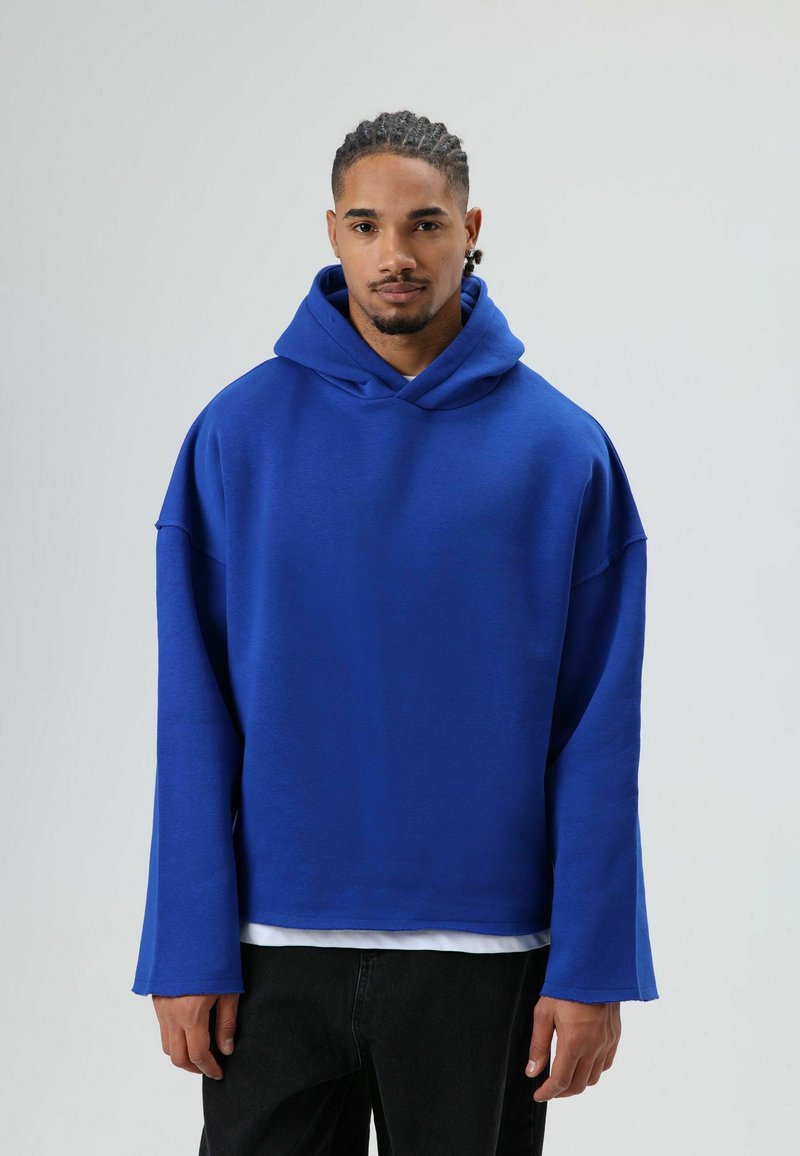 Blauwe oversized hoodie met een voorzak en een zachte textuur. Voorzien van een grote capuchon en brede mouwen, gedragen over een wit shirt.