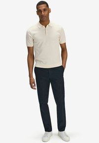 Beige kortärmad polo med en subtil textur, i kombination med marinblå byxor och vita sneakers. Modellen står med händerna i fickorna.