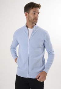 Cárdigan de cremallera azul claro con textura de canalé, cuello alto y dos bolsillos laterales, combinado con una camiseta blanca y vaqueros oscuros.