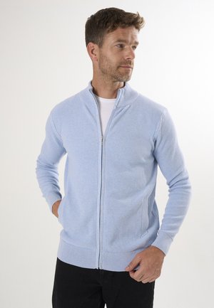 Cárdigan de cremallera azul claro con textura de canalé, cuello alto y dos bolsillos laterales, combinado con una camiseta blanca y vaqueros oscuros.