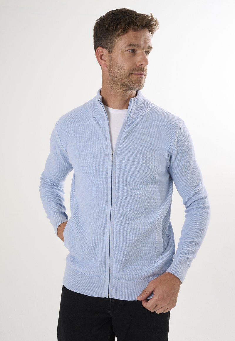 Cárdigan de cremallera azul claro con textura de canalé, cuello alto y dos bolsillos laterales, combinado con una camiseta blanca y vaqueros oscuros.