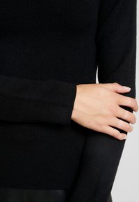 Svart ribbad långärmad tröja med mjuk textur, figurnära design och smyckade ärmar, visas med en hand som vilar på armen.